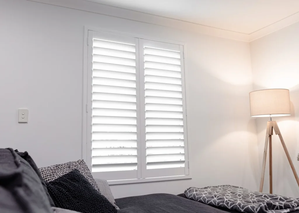 White plantation shutters on a bedroom window with a floor lamp beside a sofa