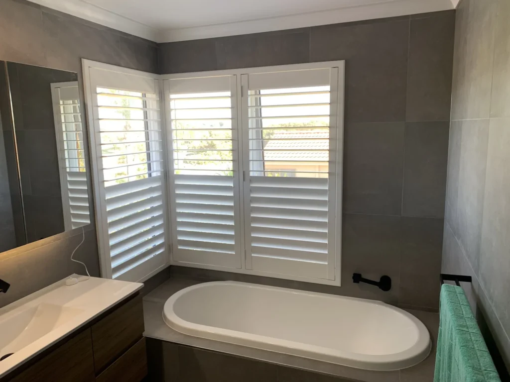 White plantation shutters fitted around a corner window above a bathtub