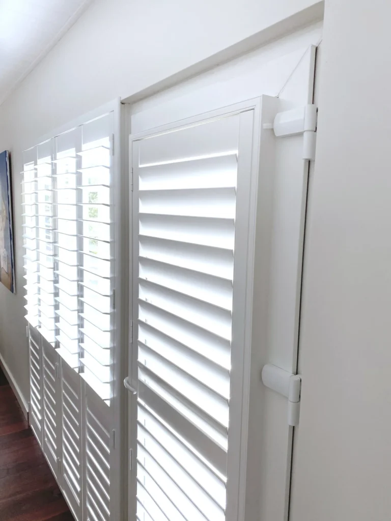 White PVC plantation shutters along a row of windows, one panel swung open